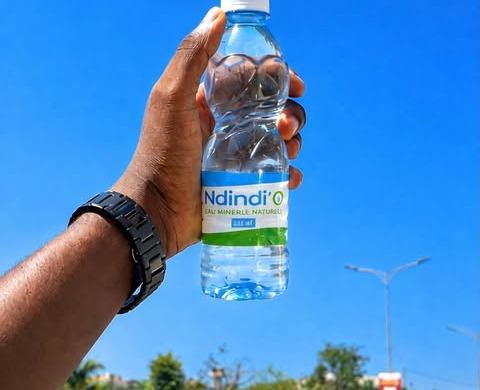 Une bouteille Ndindi'O 350 ml tenue à bout de bras sous le ciel de Touba